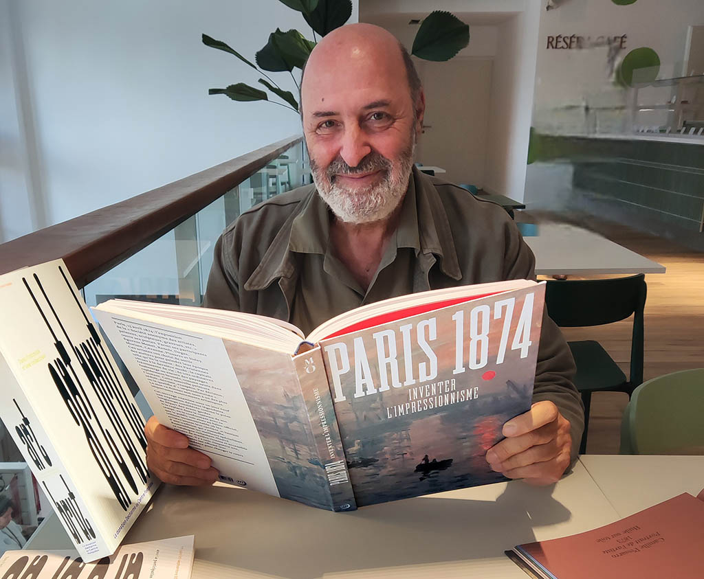 Prise de vue au sein du Grand Palais, au Réséda Café. Cédric Klapisch est assis à une table du café et fait face au photographe en tenant le catalogue ouvert de Paris 1874